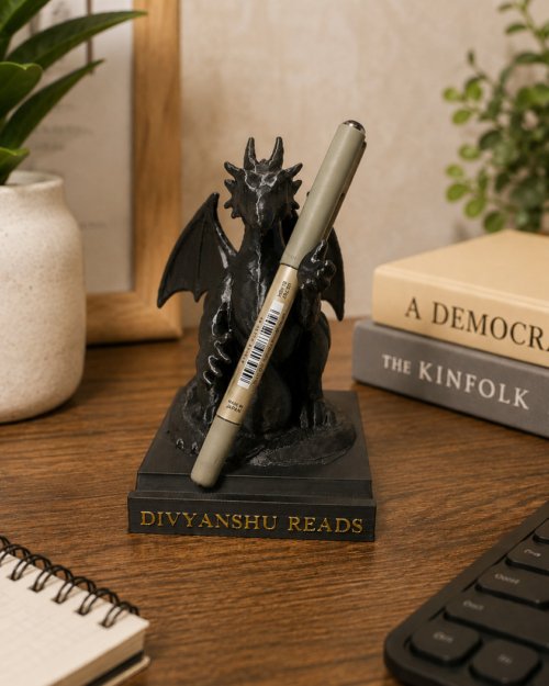 Black dragon statue holding a beige pen on its claws perched over a book labeled DIVYANSHU READS on a wooden desk, with plants and other books nearby.